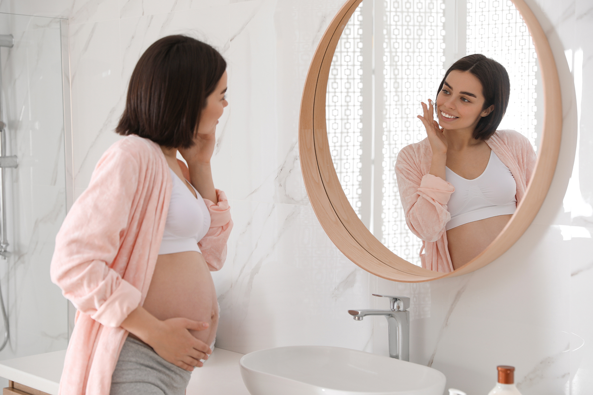 Pregnant woman in pink robe touching face and looking in mirror, considering Botox safety during pregnancy.