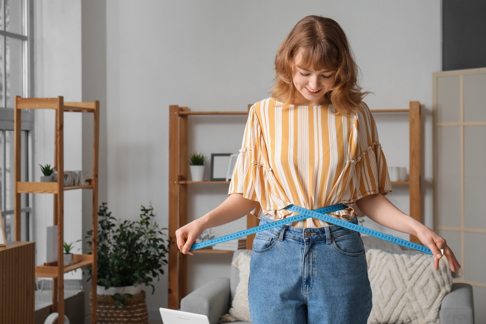Smiling woman measuring waist with tape in living room, symbolizing affordable access to semaglutide through assistance programs.
