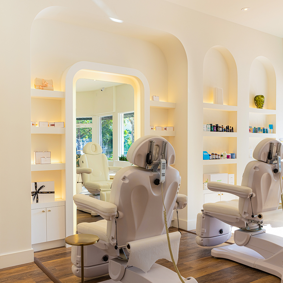 Modern treatment room at AM Beauty & Wellness in Miami with white chairs, soft lighting, and skincare products displayed.