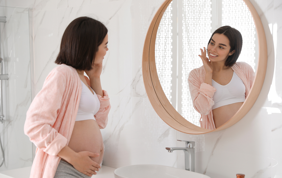 Pregnant woman in pink robe touching face and looking in mirror, considering Botox safety during pregnancy.