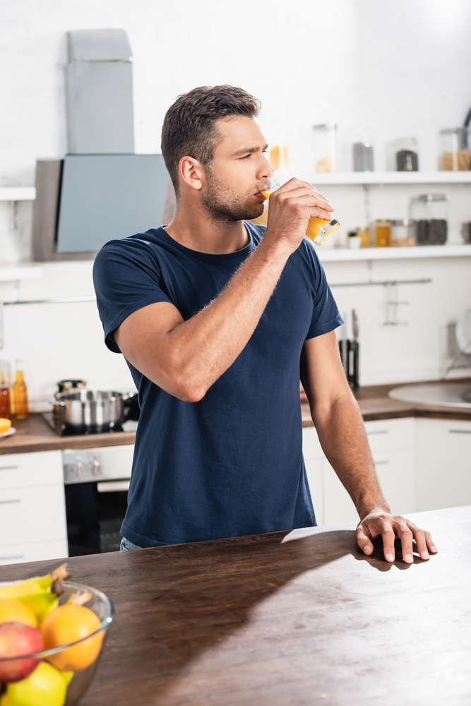 Young man in navy t-shirt sipping fresh orange juice at a bright, modern kitchen counter, healthy living concept