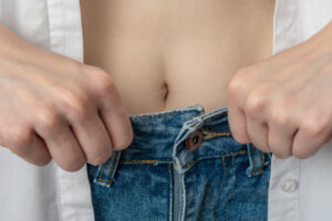 Hands struggle to button tight blue jeans, depicting weight regain after stopping Ozempic or Mounjaro treatment