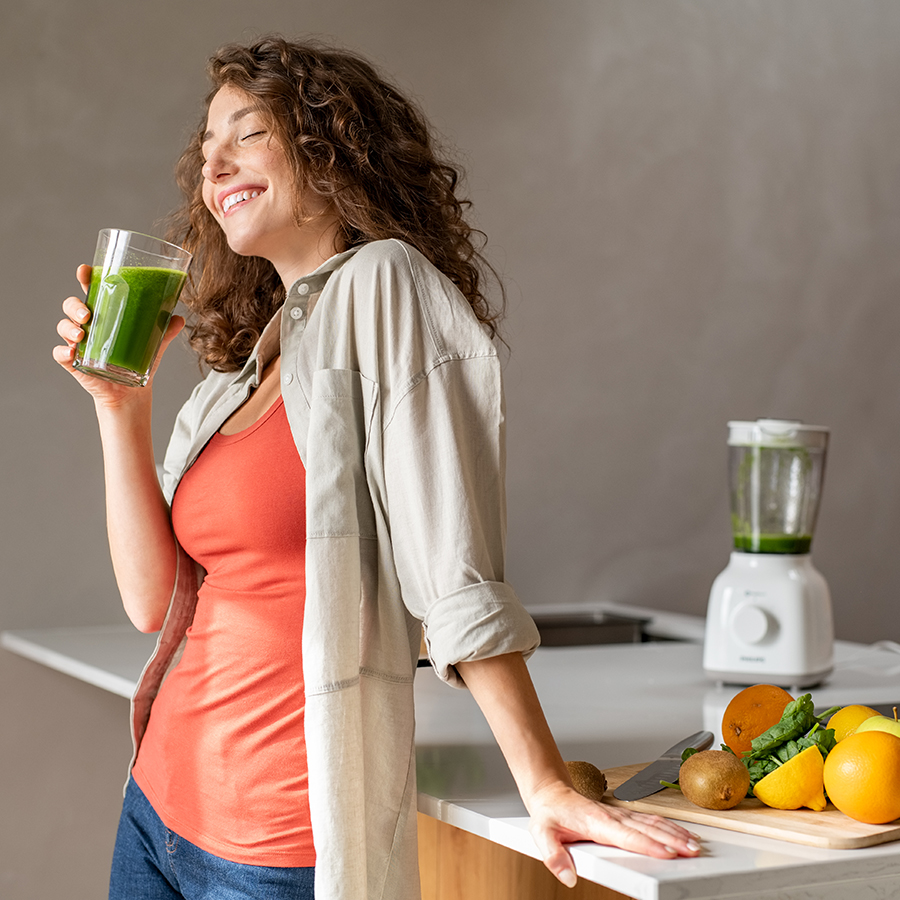 Woman enjoying green smoothie in kitchen, highlighting how lifestyle choices support hormone health and balance.