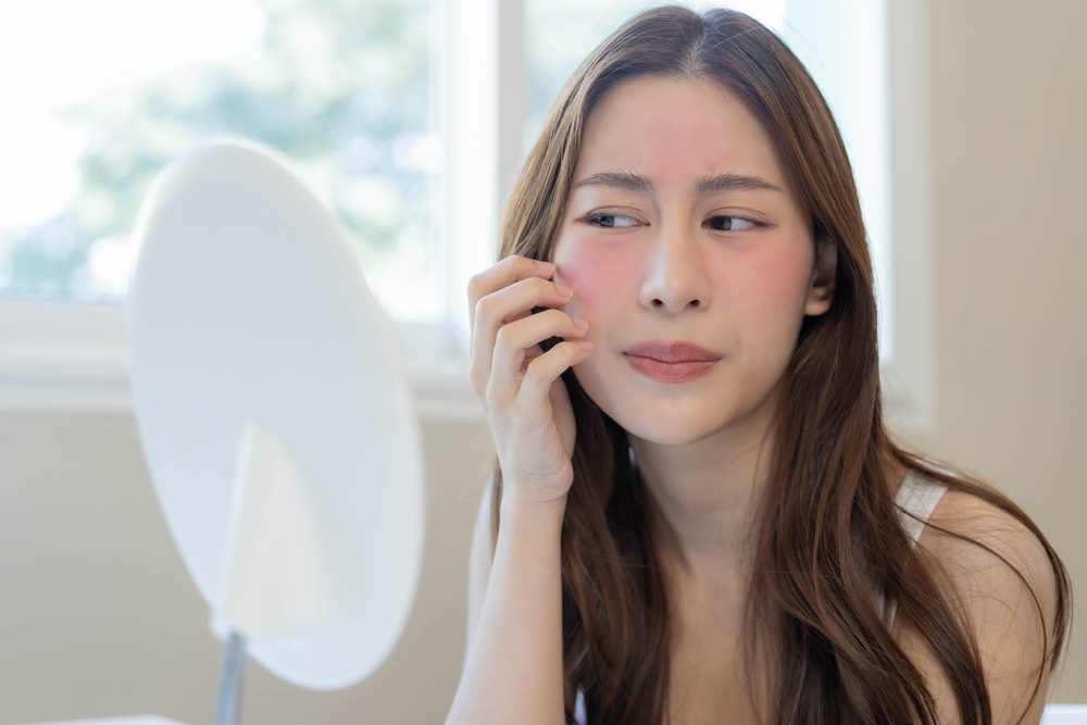 Woman examining red, sensitive skin in mirror, showing common rosacea symptoms like facial flushing and irritation.