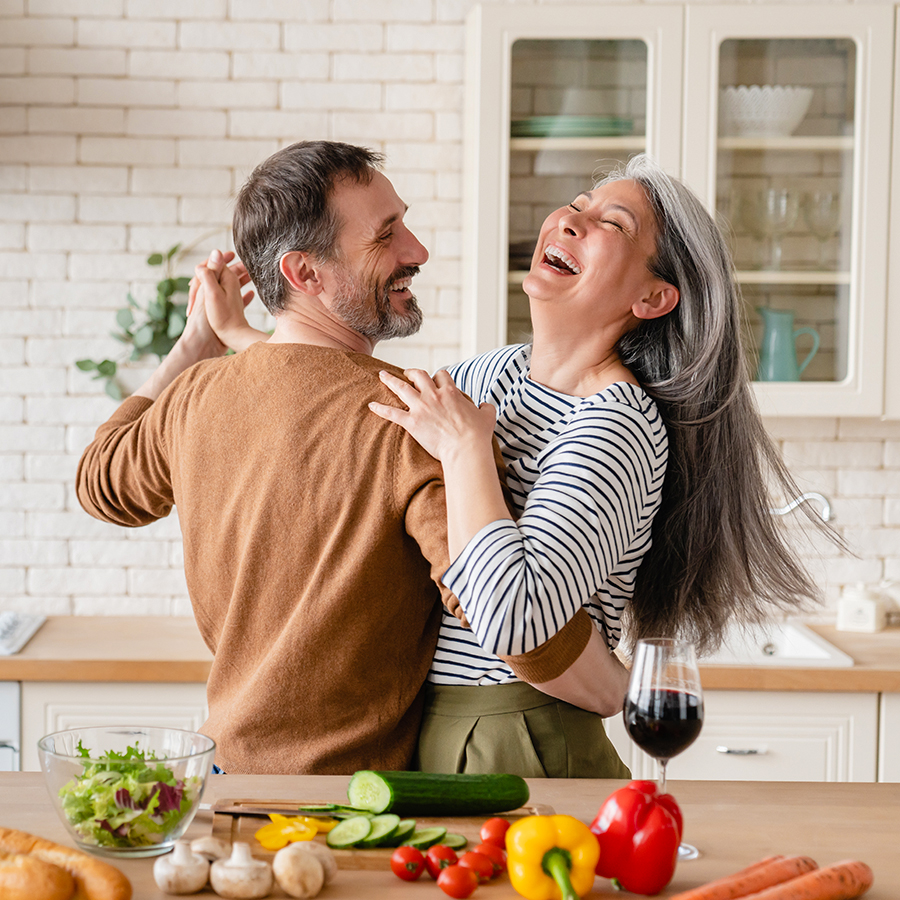 Joyful middle-aged couple dancing in the kitchen surrounded by fresh vegetables, symbolizing immune health benefits