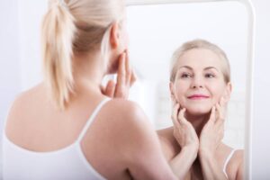 A blonde woman looking at her reflection in a mirror while gently touching her jawline and checking for fine lines.
