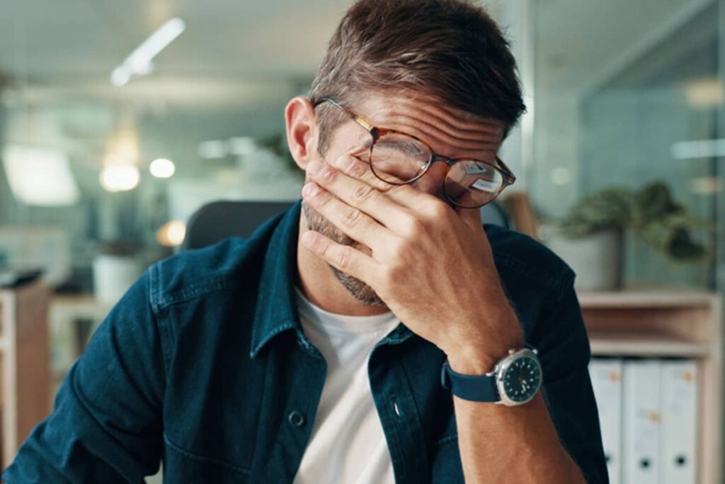 A man wearing glasses and a denim shirt rubbing his tired eyes in an office setting, illustrating symptoms of premature cellular aging.