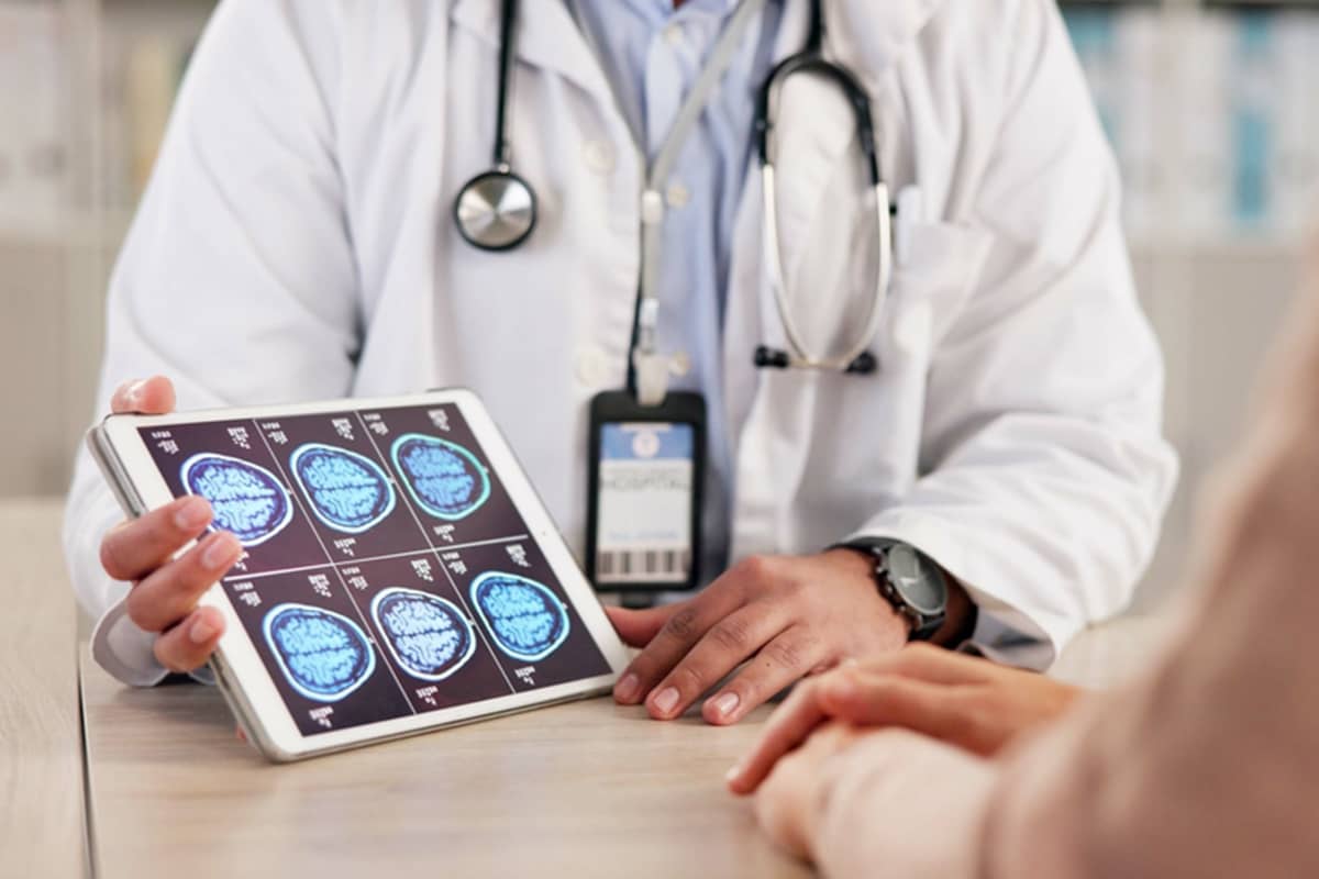 A doctor in a white lab coat holds a tablet displaying multiple blue MRI brain scans during a consultation with a patient.