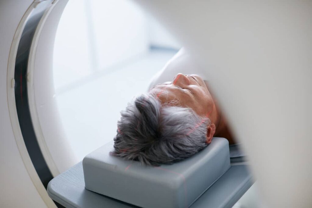 A senior man lies on a medical table with red laser alignment marks on his head as he is moved into an MRI or CT scanner.