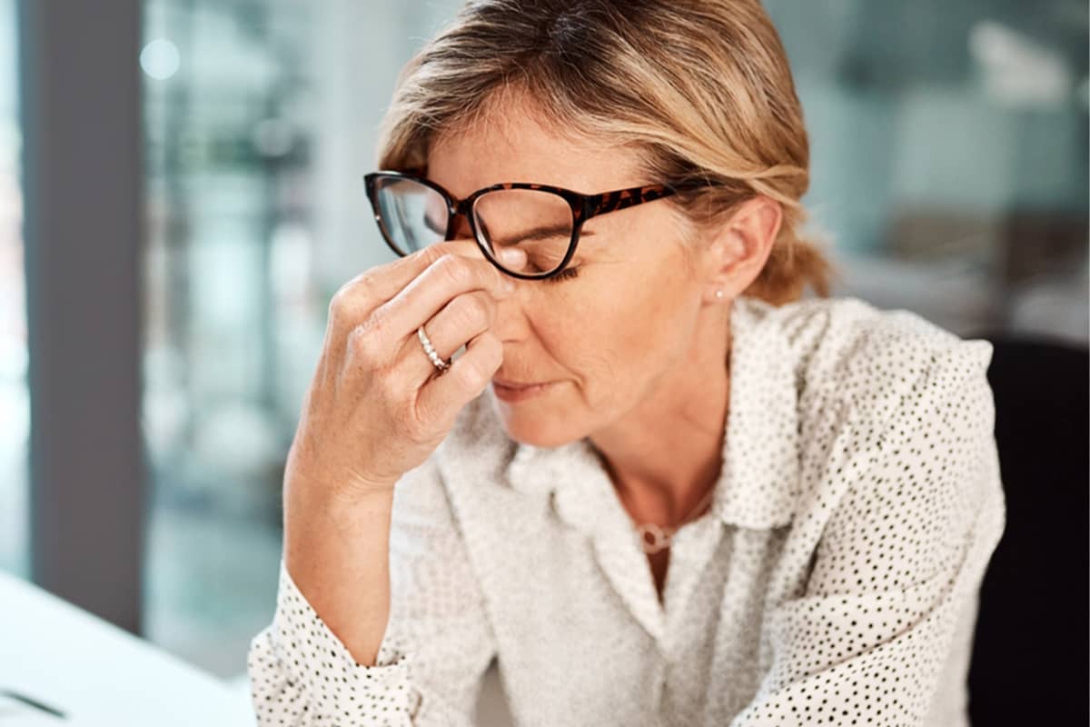 A woman wearing glasses and a polka-dot shirt pinches the bridge of her nose with her eyes closed, looking stressed and tired.