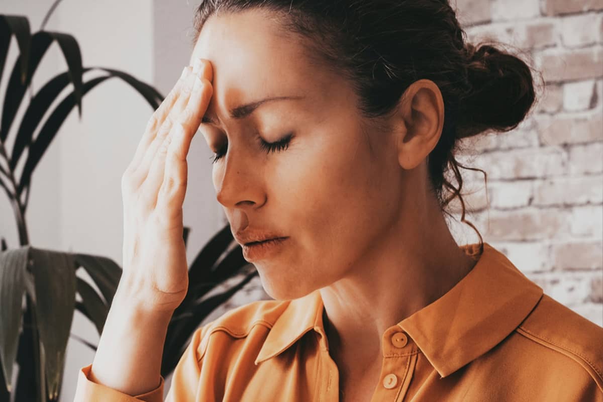 A woman in an orange shirt holds her hand to her forehead with her eyes closed, appearing to suffer from a headache or exhaustion.
