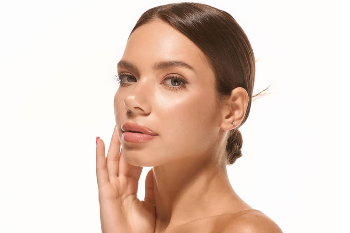 A young woman with radiant, smooth skin and a sleek hair bun posing with her hand gently touching her face to showcase clinical skin-tightening results.