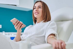 A woman in a white robe sits comfortably in a clinic chair receiving an IV infusion while holding her smartphone.