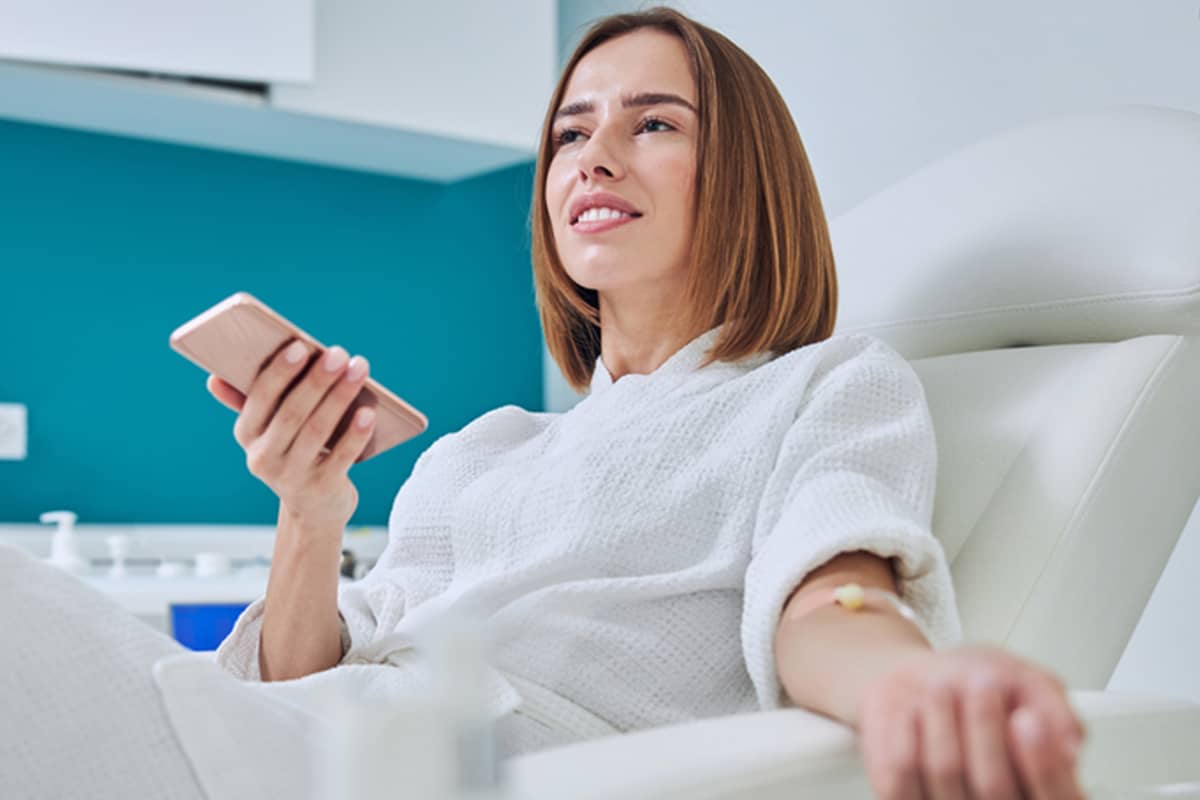 A woman in a white robe sits comfortably in a clinic chair receiving an IV infusion while holding her smartphone.