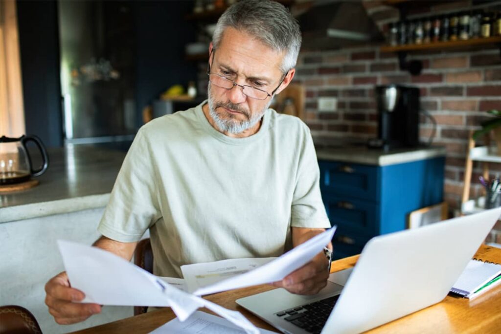 A middle-aged man with glasses and a grey beard focuses intently on paperwork and his laptop in a home kitchen setting.