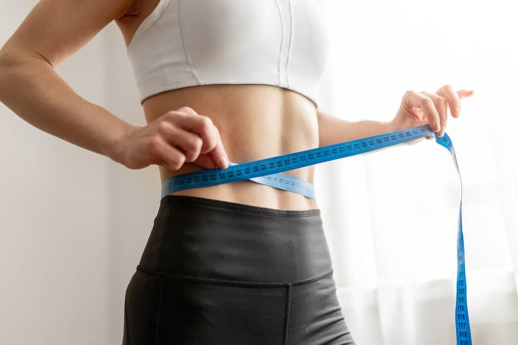 A woman in athletic wear using a blue measuring tape to track the progress of her waistline reduction.
