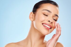 A smiling woman with smooth, radiant skin and eyes closed gently touching her cheek against a light blue background.