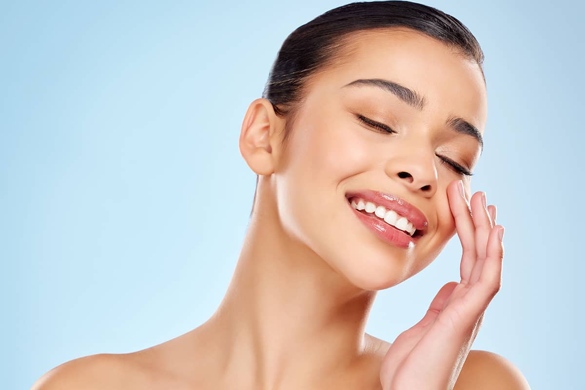 A smiling woman with smooth, radiant skin and eyes closed gently touching her cheek against a light blue background.