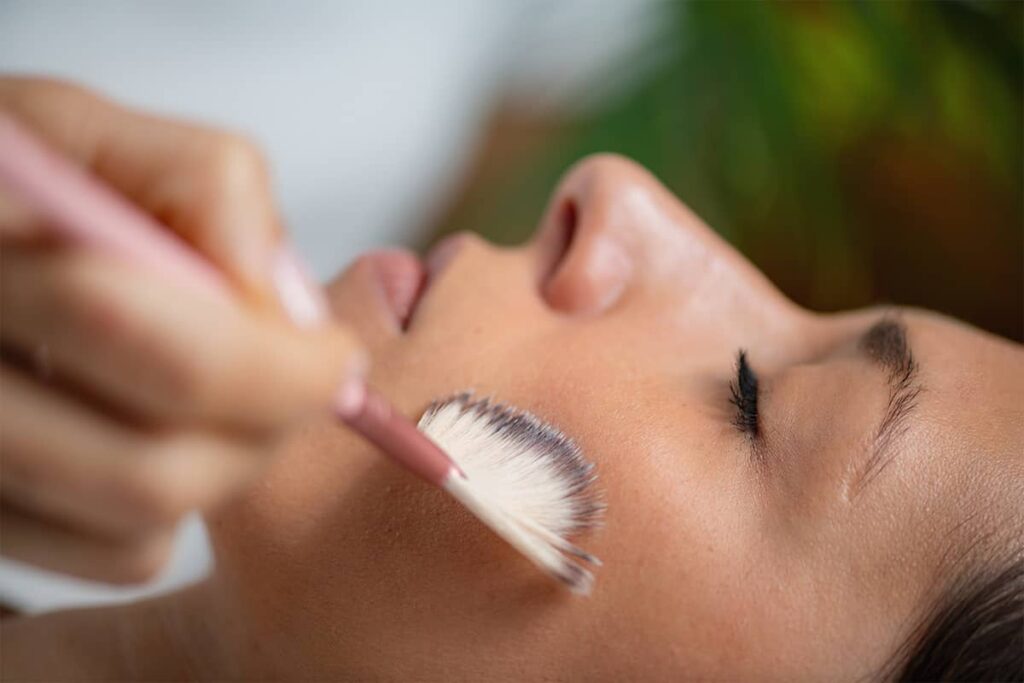 A close-up, side profile of a person receiving a chemical peel treatment as an aesthetician uses a fan brush to apply the solution to their cheek.