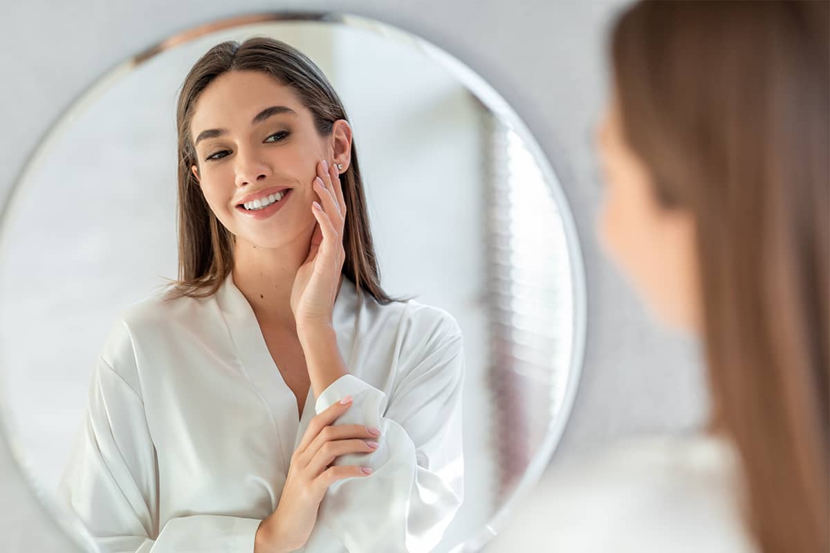 A smiling woman in a white silk robe looking at her reflection in a large circular mirror while gently touching her cheek.