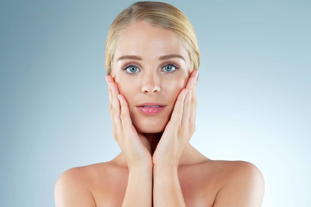 A close-up portrait of a woman with clear, glowing skin and blue eyes, holding her face with both hands against a light blue background.