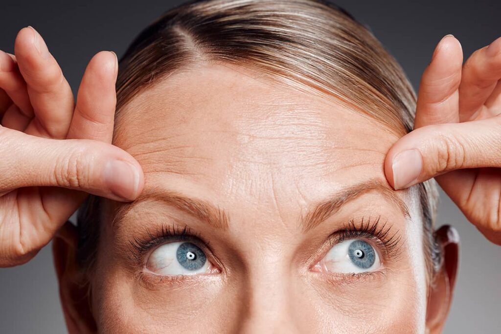 An extreme close-up of a woman's forehead showing visible horizontal wrinkles as she looks upward and touches her brow area.