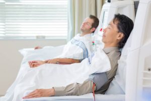 Two men resting with their eyes closed in medical chairs while undergoing an intravenous blood filtering procedure in a clinic.