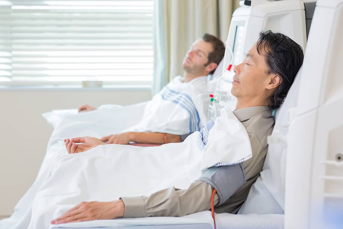 Two men resting with their eyes closed in medical chairs while undergoing an intravenous blood filtering procedure in a clinic.