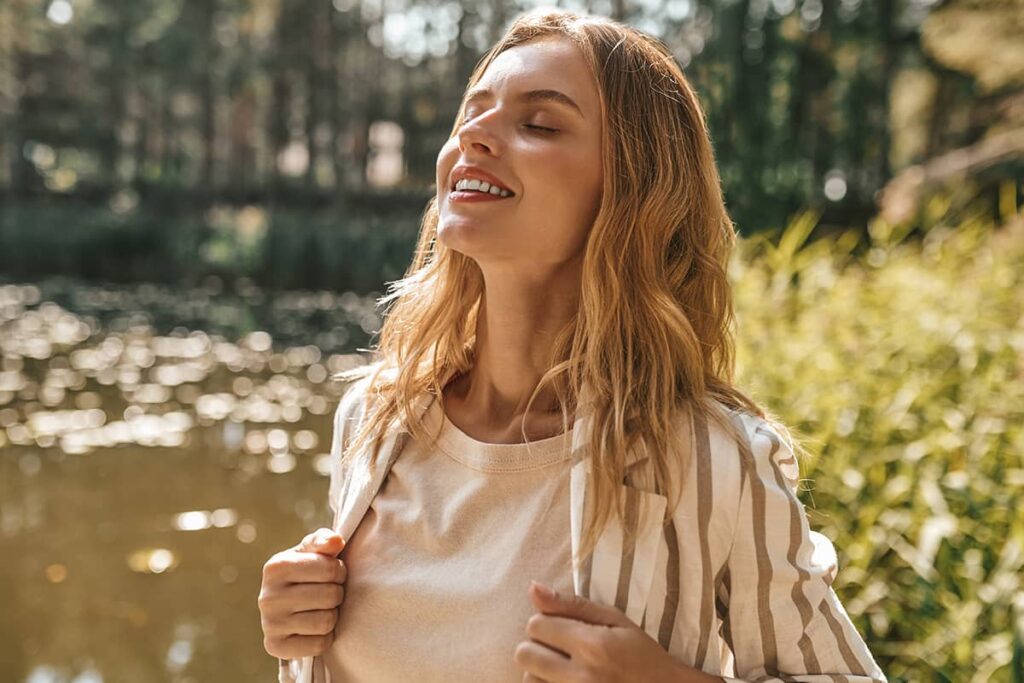 A smiling blonde woman with her eyes closed tilting her face toward the sun while standing outside near a pond.