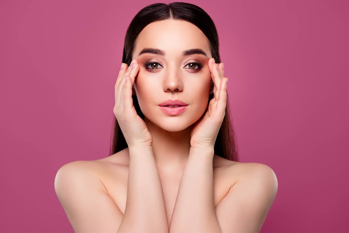 Front portrait of a brunette woman with bare shoulders gently framing her face with both hands against a solid pink background.