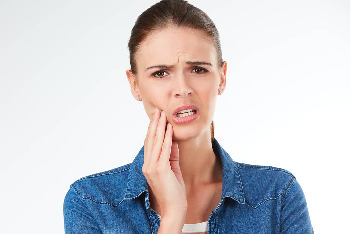 A woman in a blue denim shirt looking forward with a concerned expression while holding her hand against her lower cheek and jaw.