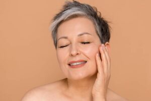 A close-up portrait of a mature woman with short, stylish gray hair smiling with her eyes closed while gently resting her fingertips against her cheek.