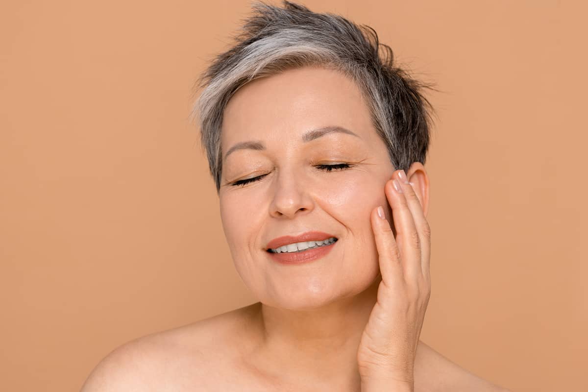 A close-up portrait of a mature woman with short, stylish gray hair smiling with her eyes closed while gently resting her fingertips against her cheek.