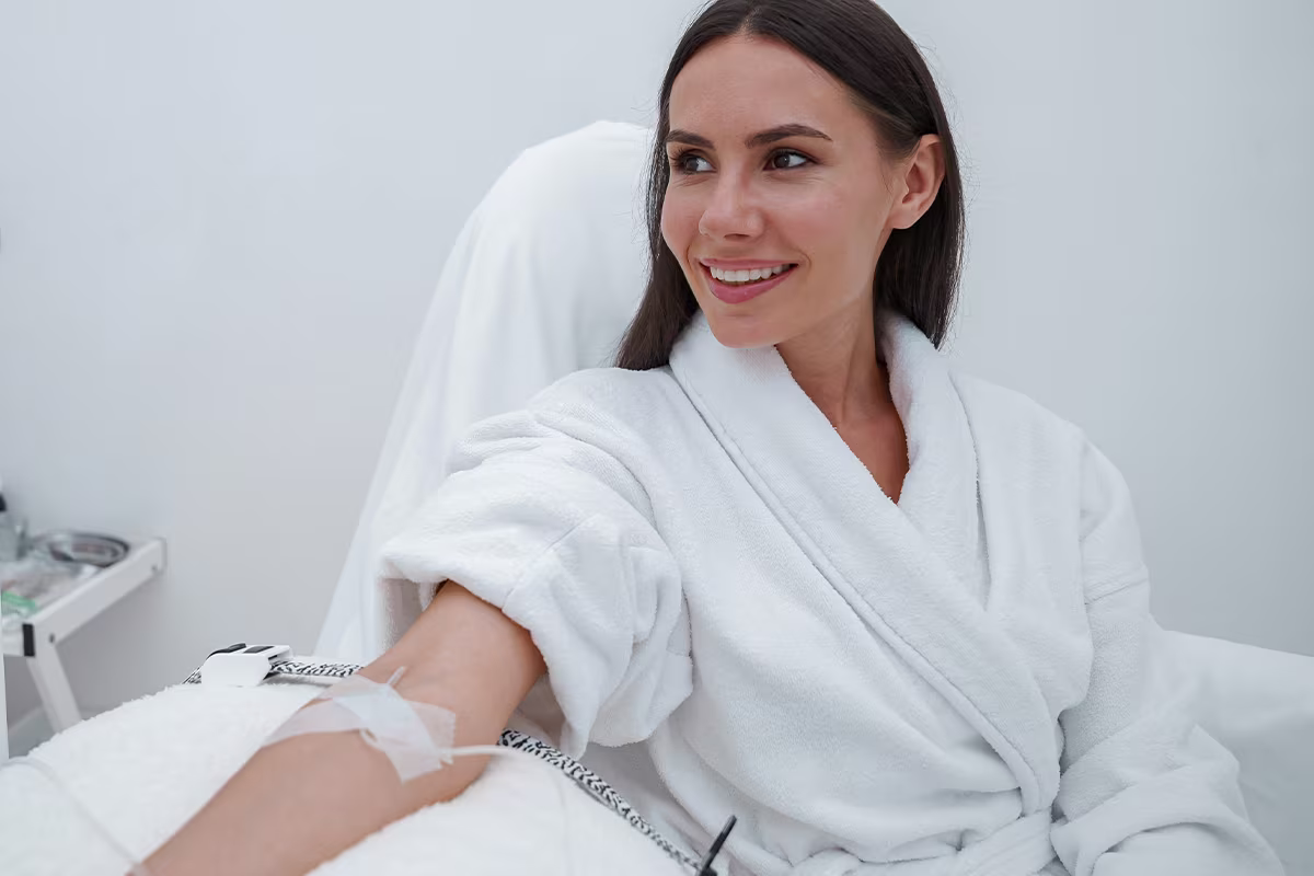 A smiling woman in a white robe receiving intravenous therapy in a bright clinical room.