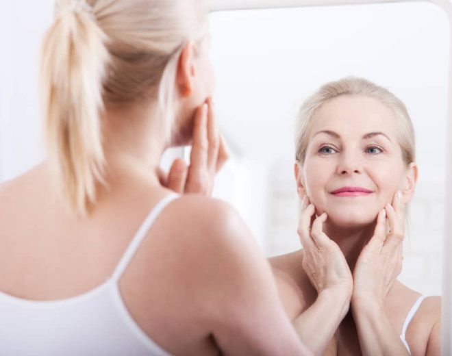 A blonde woman looking at her reflection in a mirror while gently touching her jawline and checking for fine lines.