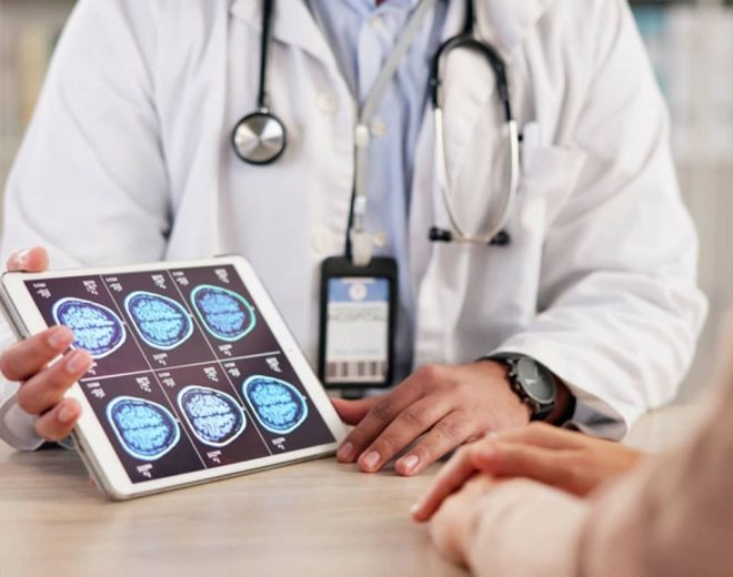 A doctor in a white lab coat holds a tablet displaying multiple blue MRI brain scans during a consultation with a patient.