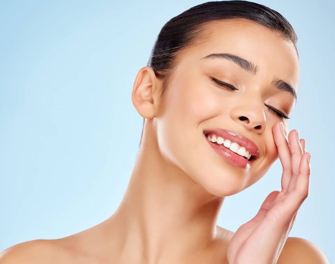A smiling woman with smooth, radiant skin and eyes closed gently touching her cheek against a light blue background.