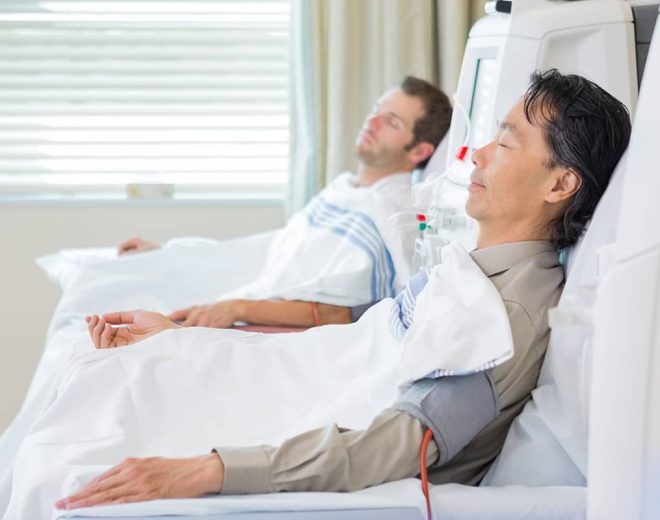 Two men resting with their eyes closed in medical chairs while undergoing an intravenous blood filtering procedure in a clinic.