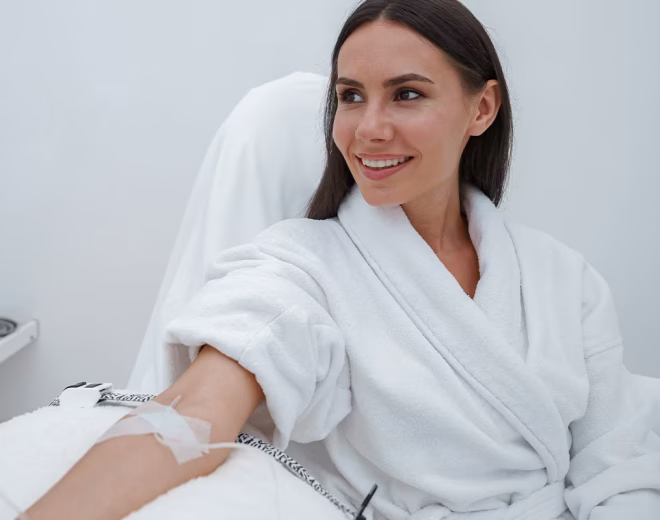 A smiling woman in a white robe receiving intravenous therapy in a bright clinical room.