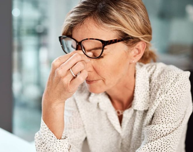 A woman wearing glasses and a polka-dot shirt pinches the bridge of her nose with her eyes closed, looking stressed and tired.