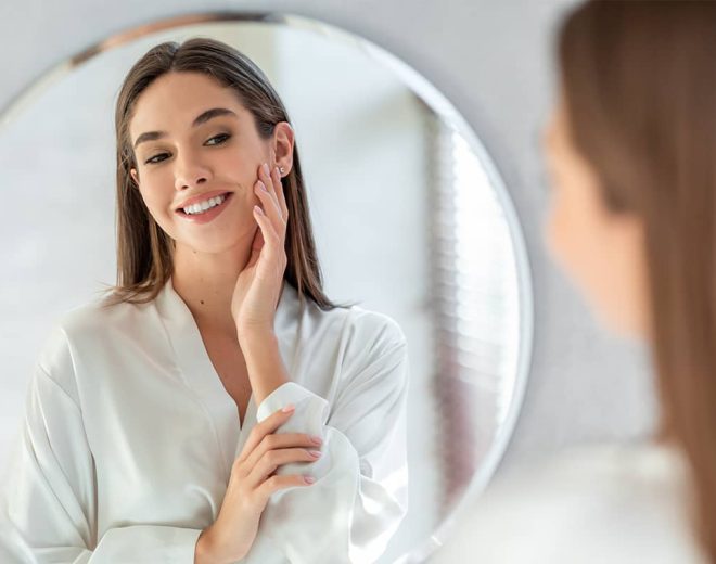 A smiling woman in a white silk robe looking at her reflection in a large circular mirror while gently touching her cheek.