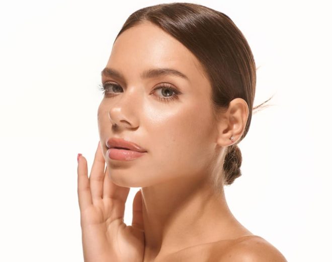A young woman with radiant, smooth skin and a sleek hair bun posing with her hand gently touching her face to showcase clinical skin-tightening results.