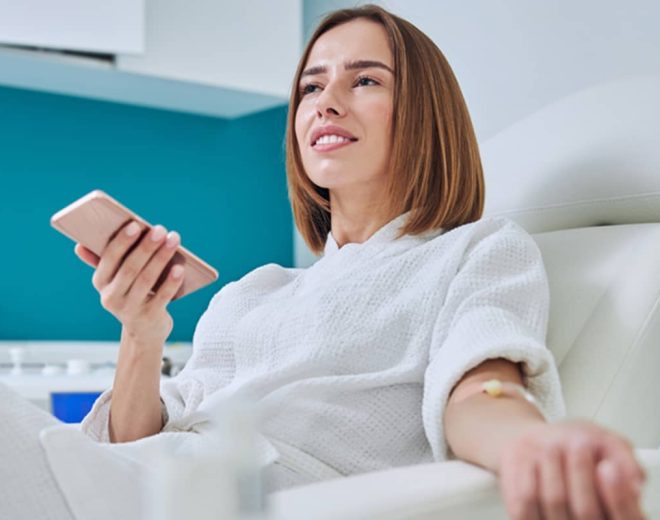 A woman in a white robe sits comfortably in a clinic chair receiving an IV infusion while holding her smartphone.