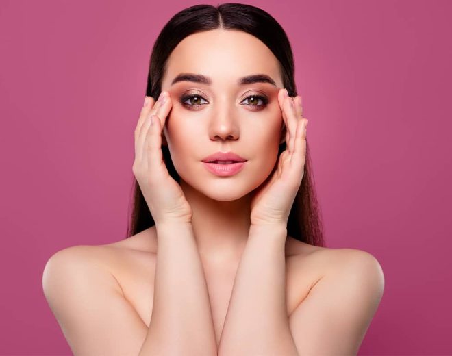 Front portrait of a brunette woman with bare shoulders gently framing her face with both hands against a solid pink background.