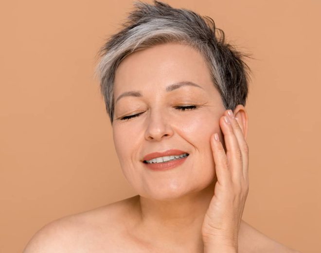 A close-up portrait of a mature woman with short, stylish gray hair smiling with her eyes closed while gently resting her fingertips against her cheek.